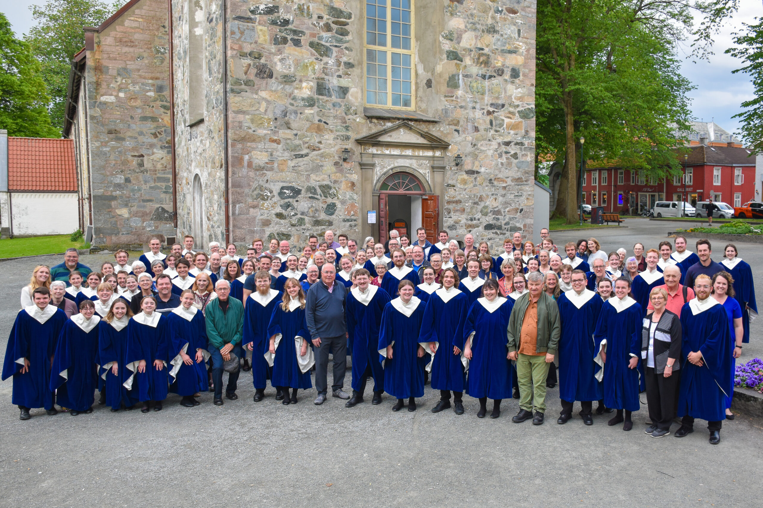 A large group of people, most in blue choir robes, stand in front of an old, large stone building