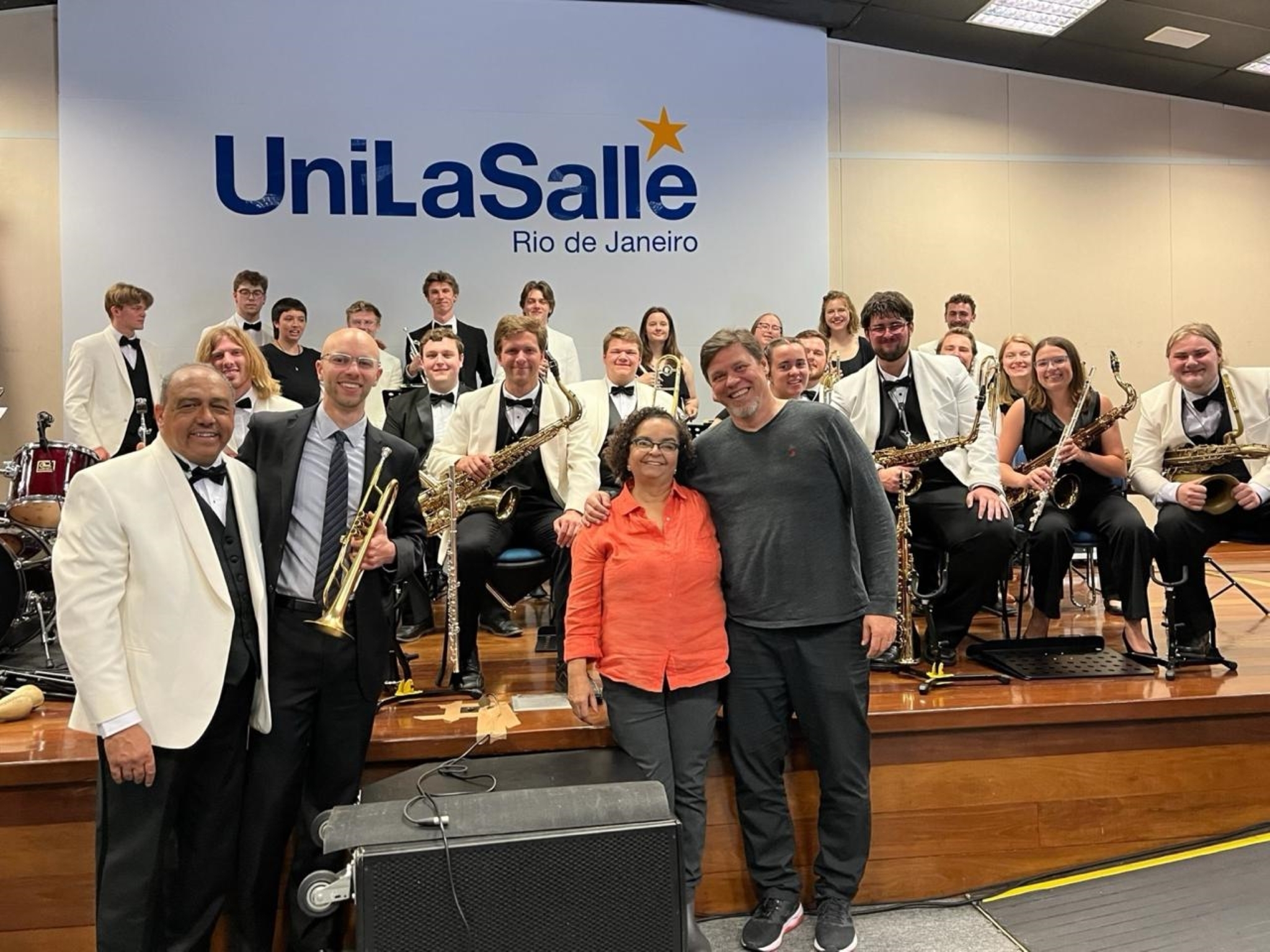 A jazz orchestra, some holding brass instruments, sits in front of a banner