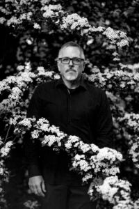 Black and white phots of man in dark top, black rimmed glasses, standing behind a branch of blooming flowers.