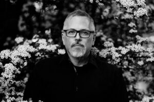 Black and white phots of man in dark top, black rimmed glasses, standing behind a branch of blooming flowers.