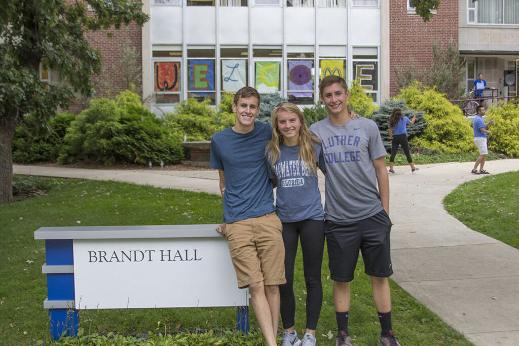 3 students rest arm in arm on the Brandt Hall sign in front of the hall, where signs in the windows spell out "Welcome"