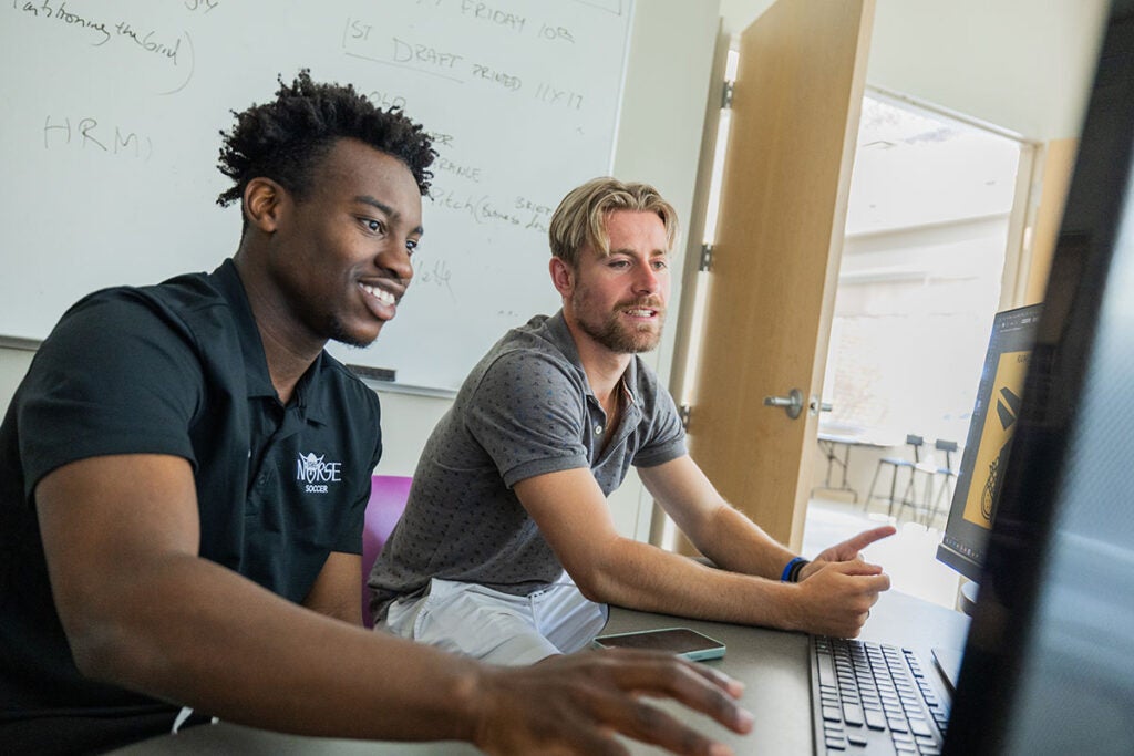Two students sit together at a desk looking at a computer