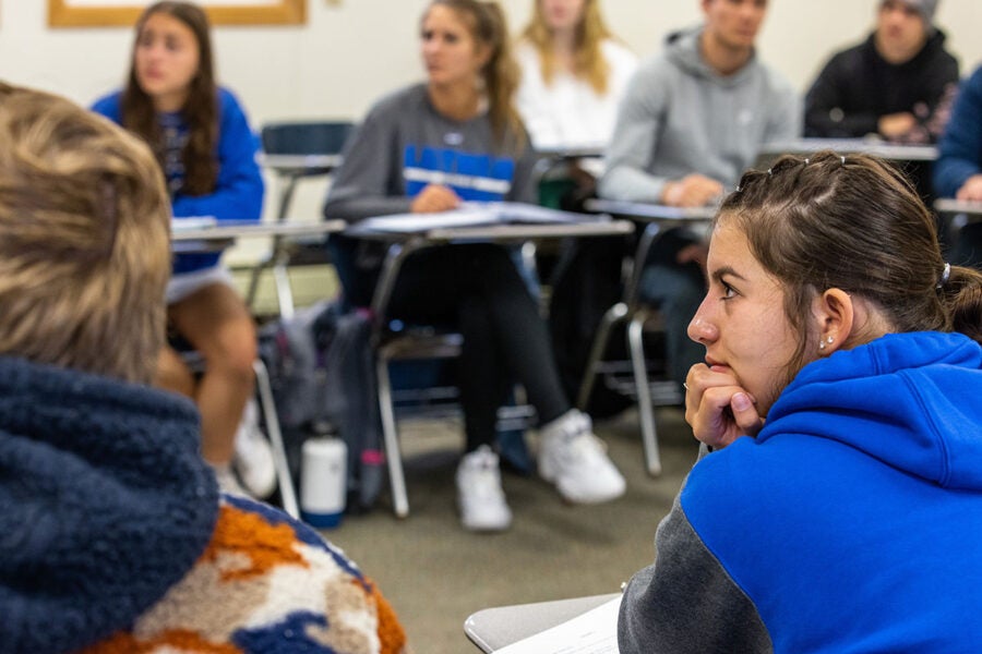 One student is shown in sharp focus from behind, sitting at a desk in a classroom.