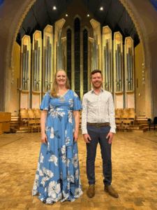 Photo of Lara Loge and Steven Luksan standing in front of a large, pip organ.