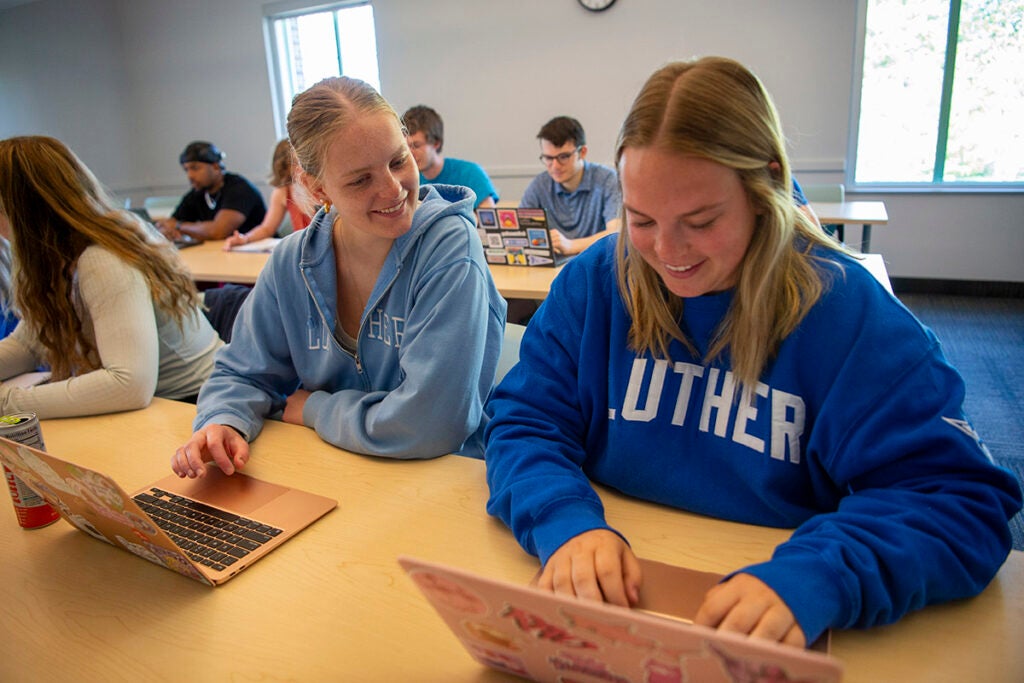 Two students sit at a table together working on laptops