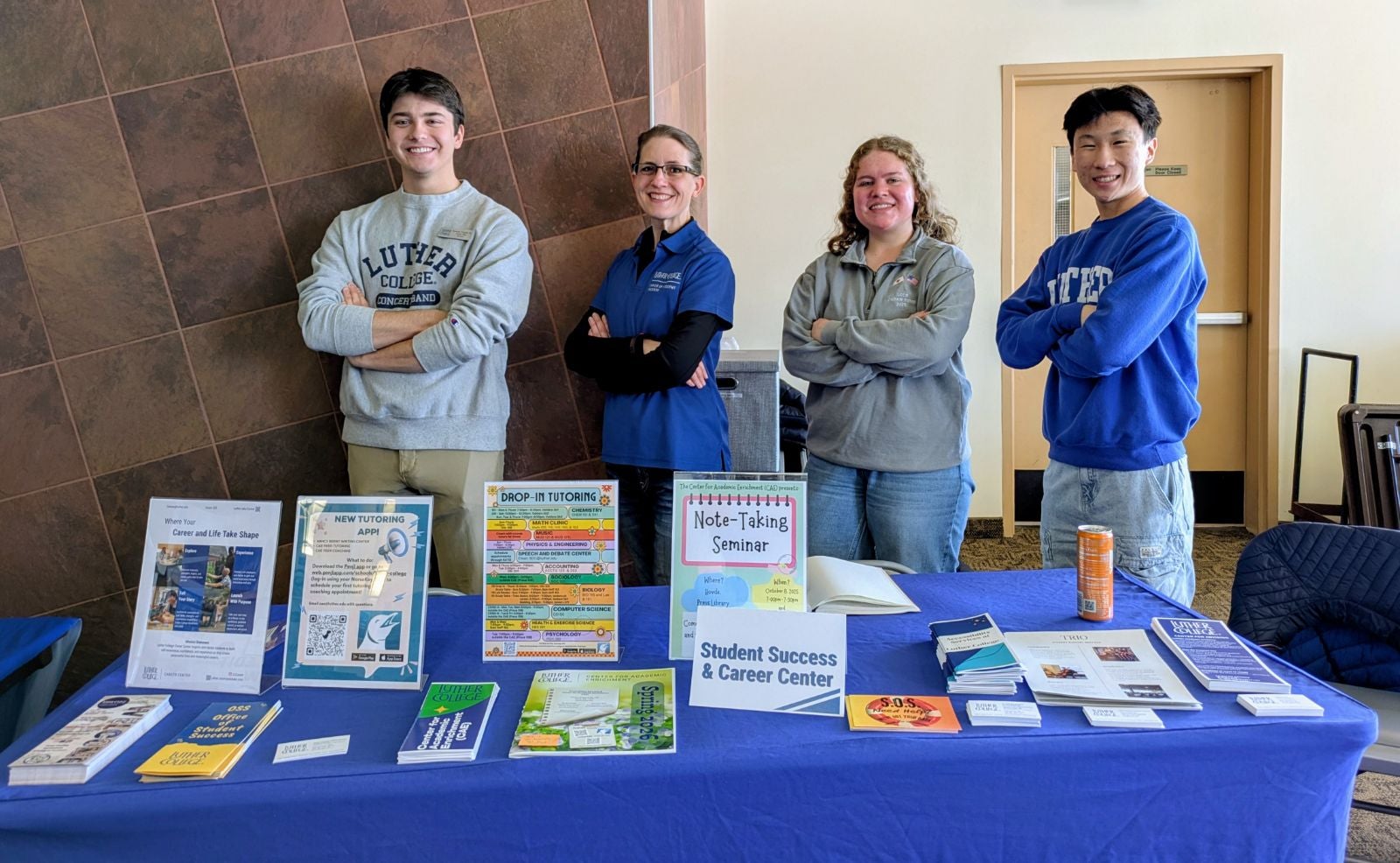 Four representatives of the Office of Student Success posing behind a blue table full of resources at an event for new students.