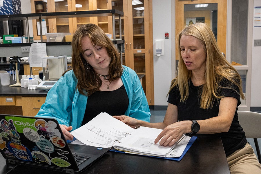 A student and a professor study a notebook with lab results.