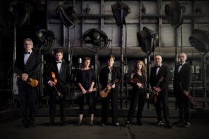 seven ukulele musicians stand for a promotional photo inside a dark room
