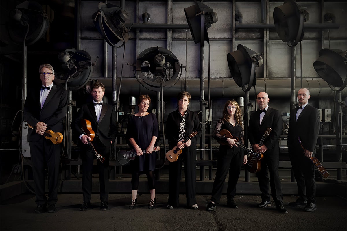 seven ukulele musicians stand for a promotional photo inside a dark room