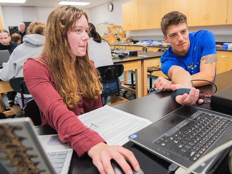 two students work together, one at a computer and the other wearing a monitor on his hand