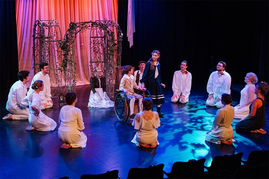 A dramatically lit stage upon which a group of about a dozen people sit in a circle around a girl standing in a Victorian navy dress and a boy in a wheelchair in Victorian-style clothing