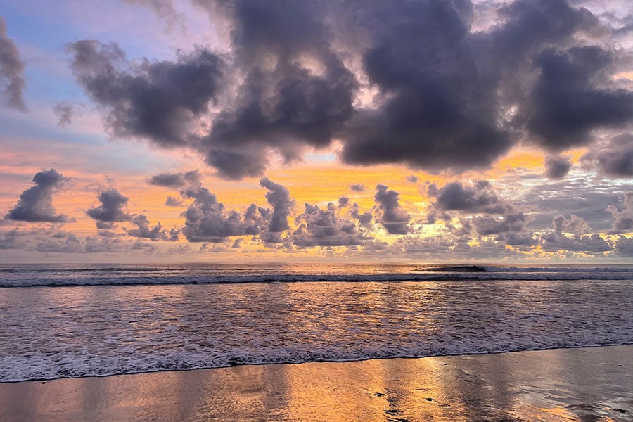 A colorful sunset of purples and oranges with many low-hanging clouds along a large ocean beach