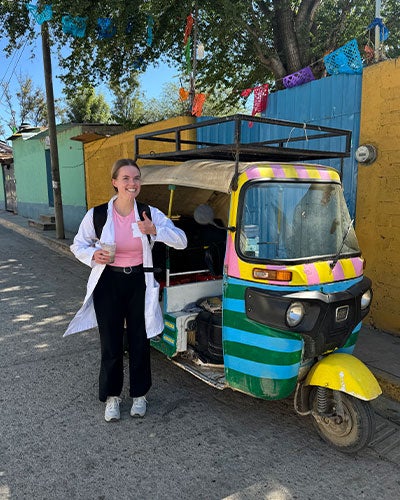 A young woman in a white medical jacket, wearing a backpack, giving a thumbs-up next to a small colorful vehicle