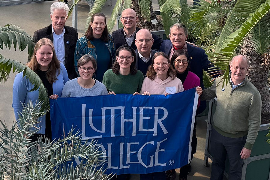 A group of 11 people in a greenhouse holding a blue Luther College banner, surrounding by palm foliage
