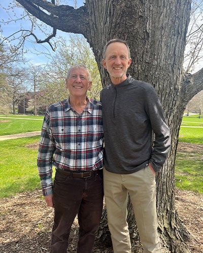 Two men standing with an arm behind each other's backs in front of a large tree