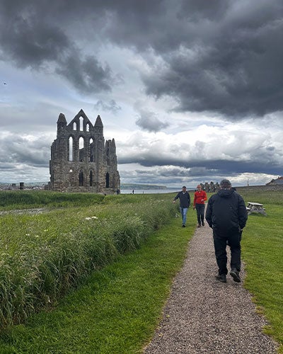 In the background are the ruins of an abbey against a cloudy gray sky. In the foreground is long grass with a trail through it and people, with their backs to the camera, walking along the trail toward the ruins.