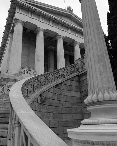 A large gray stone building with multiple columns at its front. A set of stairs with a big curved handrail leads up the building.