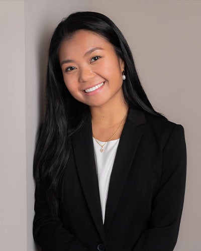 A smiling woman in a black suit jacket leaning against a beige wall