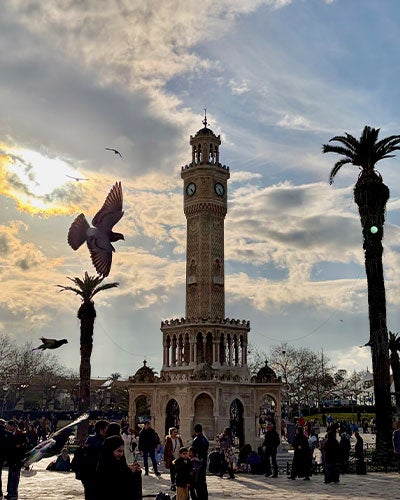 A town square with a tall clocktower in the center and two tall palm trees on either side. A crowd of people is milling around at the base of the clocktower, and a bird in flight is in next to the clocktower.