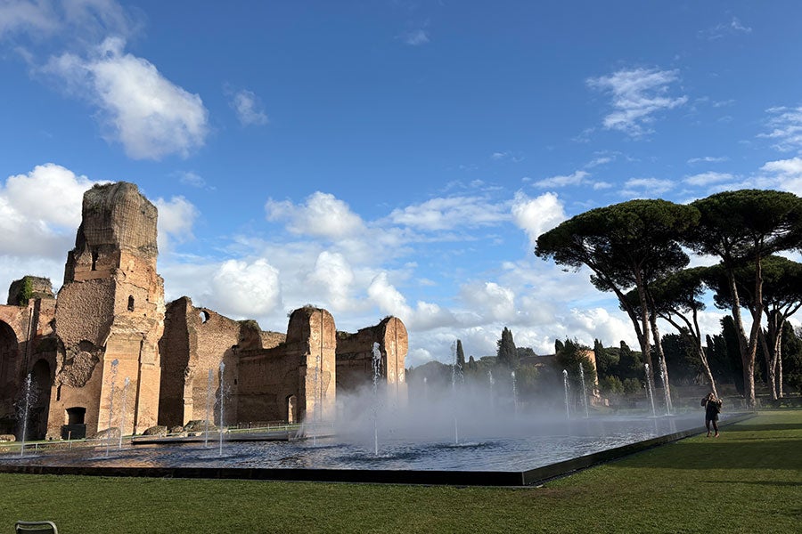 Ancient Roman ruins on the left rise up through mist. A stand of tall trees stands on the right. There is a blue sky with white clouds in the background.