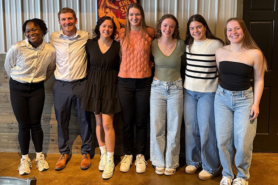 A group of seven smiling people in mostly casual clothing with their arms behind each other's backs