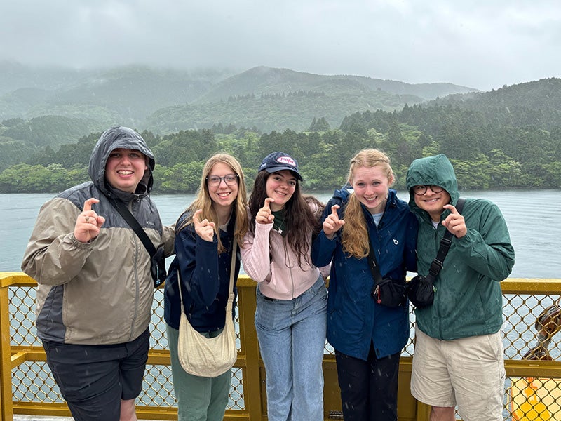 Five students on a boat deck, with a rainy background and green hills