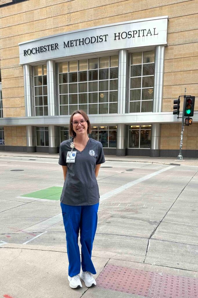 Nursing major Nora Tresemer stands in nursing scrubs in front of Rochester Methodist Hospital.