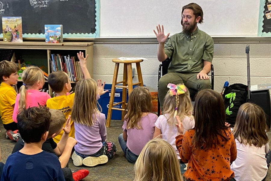 A young man teaches a class full of children, who are raising their hands.