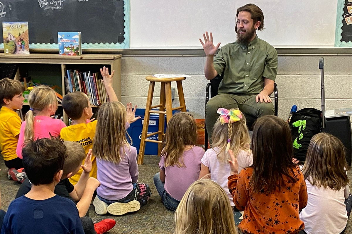 A young man teaches a class full of children, who are raising their hands.