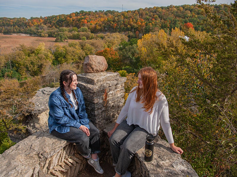 two students sit facing each other on a stone wall, overlooking the prairie and bluffs with fall foliage