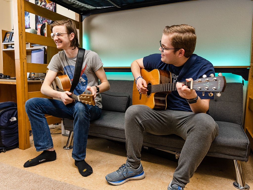 Two students sit on a couch underneath a bunked bed, playing guitars and smiling.