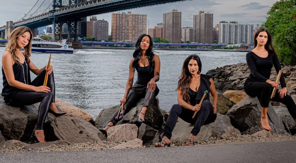The Four members of Excelsis Percussion all wearing black sitting by a river in an urban setting