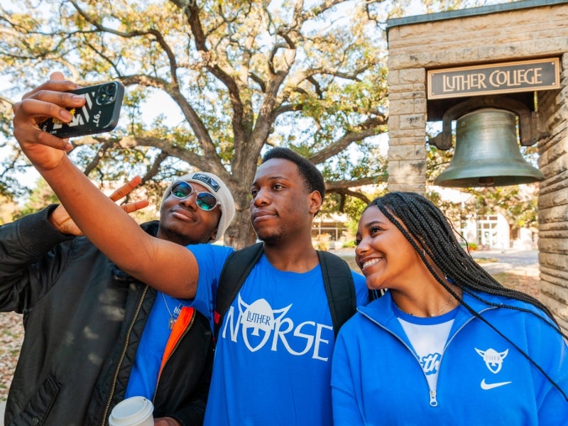 Three students pose for a selfie with the Luther College bell behind them.
