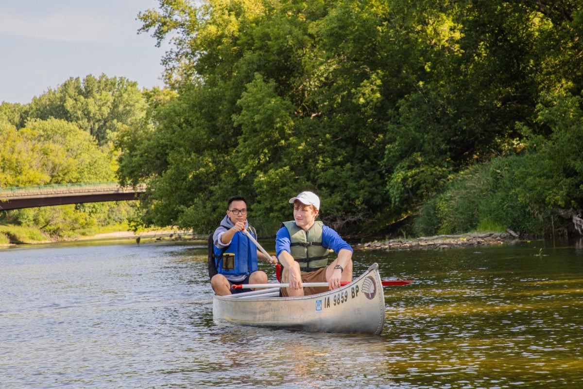 Two students paddle in a canoe on a river with a bridge and woods in the background.