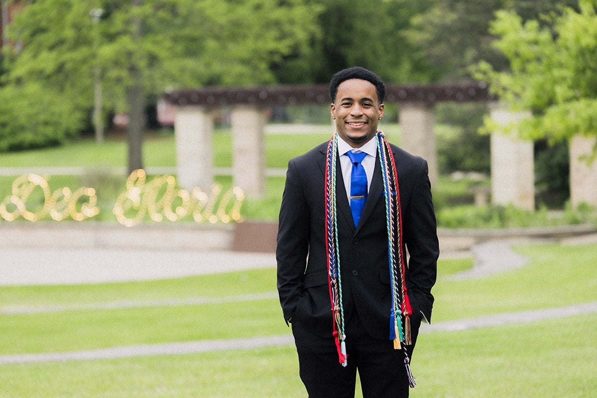 Malachi poses in a suit on campus, wearing his graduation cords.