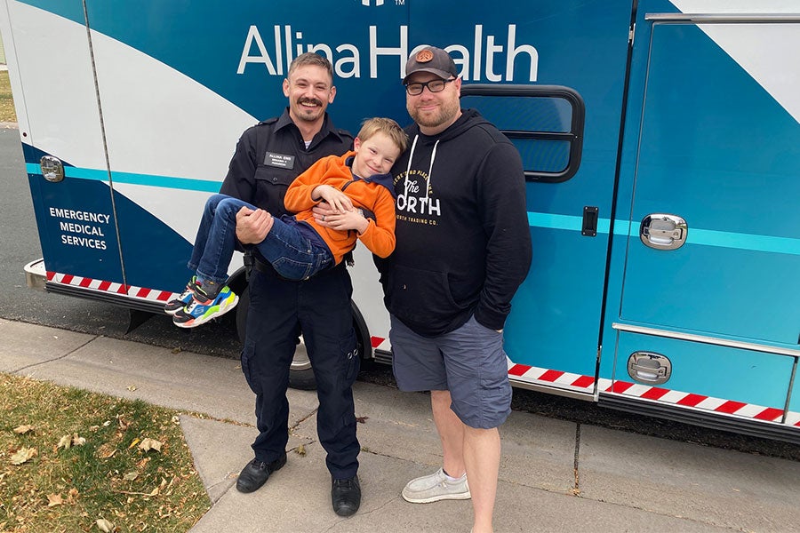 Two men stand in front of an ambulance. The man on the left wears an EMS uniform and holds a boy.