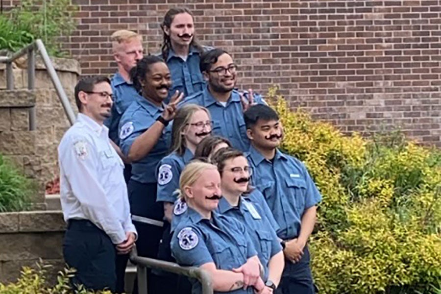 Ten people in EMS uniforms stand on outdoor stairs with fake mustaches