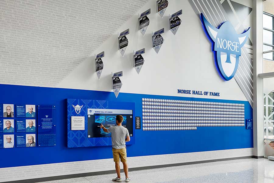 A man stands at a touchscreen on a tall wall featuring plaques and photos