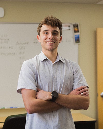 A smiling young man with his arms crossed standing in front of a whiteboard