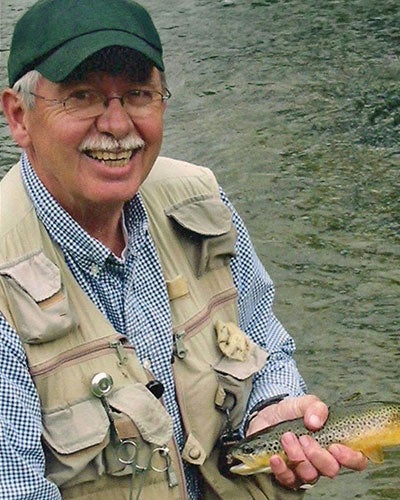 A man in a baseball hat and fishing vest holds a trout with a stream in the background