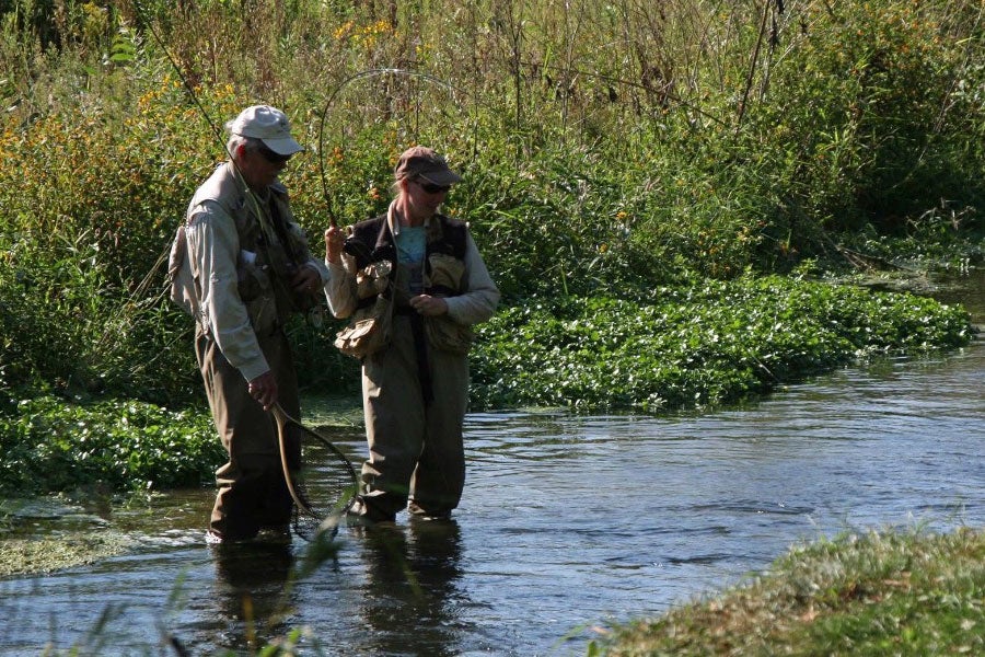 A man and woman dressed in gaiters and fishing vests stand ankle-deep in a stream. The woman holds a fishing pole.