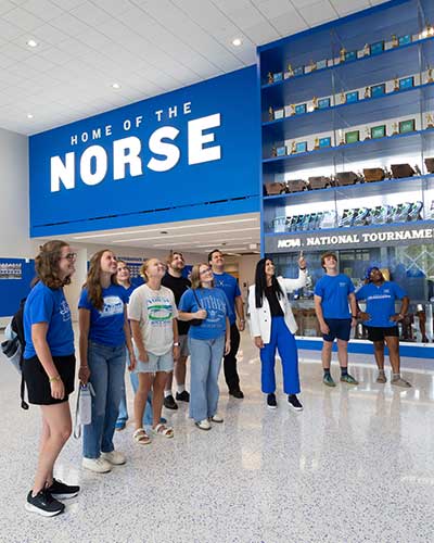 About 10 people stand in a semicircle in an expansive lobby with a wall of trophies behind them