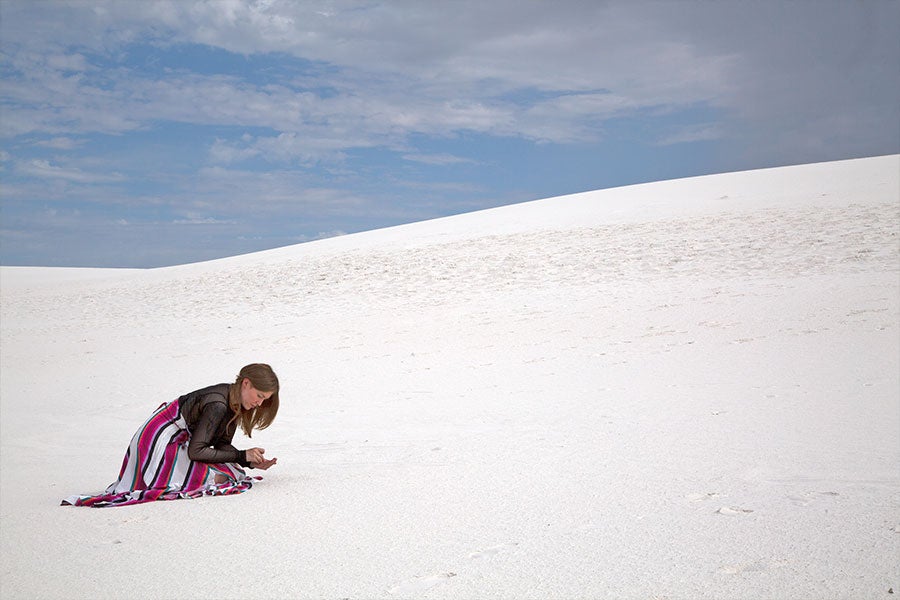 A woman in a striped skirt and black top kneels on a white sand dune, inspecting something in her hands