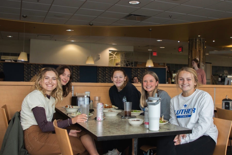 A group of students posing for a picture at the Caf, sitting at a table with their drinks and meals.