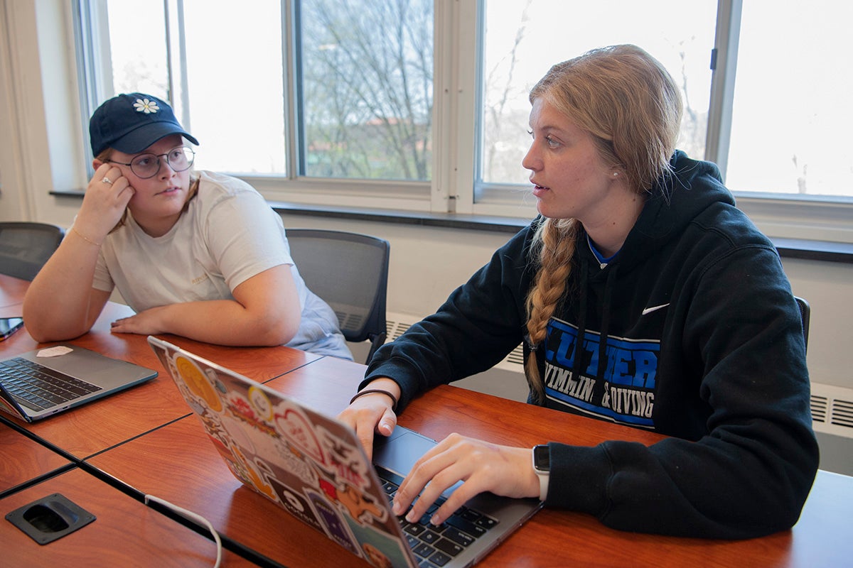 Two students sit together at a table in a classroom, talking and typing on a laptop.